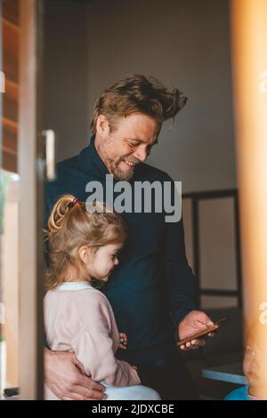 L'uomo felice che mostra il telefono cellulare alla figlia visto attraverso la finestra Foto Stock