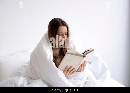 Giovane donna sorridente avvolta in coperta bianca lettura libro sul letto a casa Foto Stock