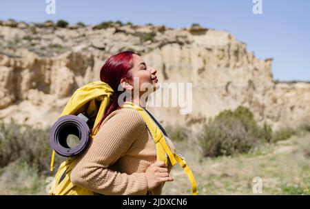 Donna sorridente che indossa uno zaino in piedi con gli occhi chiusi il giorno di sole Foto Stock