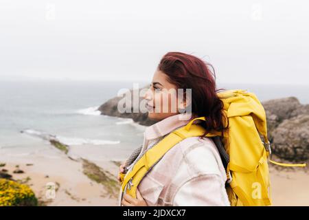 Donna sorridente con capelli rossi e zaino in piedi di fronte al mare Foto Stock