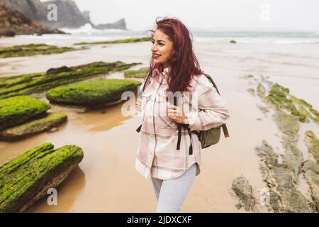 Donna sorridente con capelli rossi e zaino in piedi in spiaggia Foto Stock