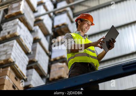 Sorridente lavoratore che indossa l'hardhat utilizzando un tablet PC da ringhiera in magazzino Foto Stock