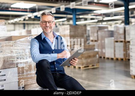 Buon manager che indossa occhiali con tablet PC seduto di fronte alla scatola di cartone in magazzino Foto Stock