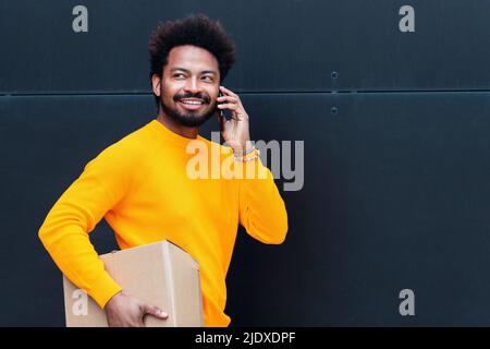 Uomo di consegna sorridente che tiene la scatola che parla sullo smartphone di fronte alla parete nera Foto Stock