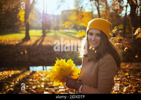 Ciao ottobre. Felice donna moderna di 40 anni in cappotto marrone e cappello giallo con foglie di giallo autunno all'aperto sul parco della città in autunno. Foto Stock