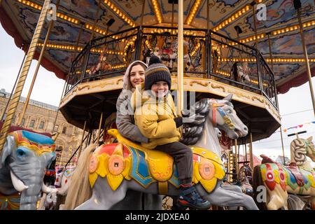 Ragazzo sorridente su cavallo carosello da madre nel mercato di Natale Foto Stock