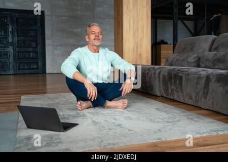 Uomo con gli occhi chiusi che fa yoga da computer portatile su tappeto a casa Foto Stock