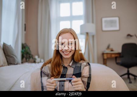 Donna felice redhead che indossa gli occhiali seduti con il test di gravidanza di fronte al letto a casa Foto Stock