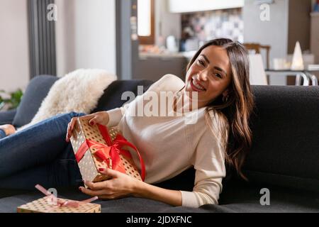 Felice bella donna con regalo di Natale reclinata sul divano in soggiorno Foto Stock