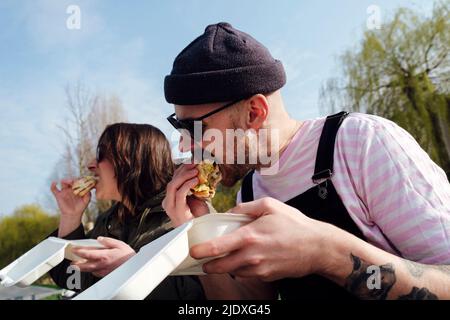 Amici affamati che mangiano hamburger nel parco il giorno di sole Foto Stock