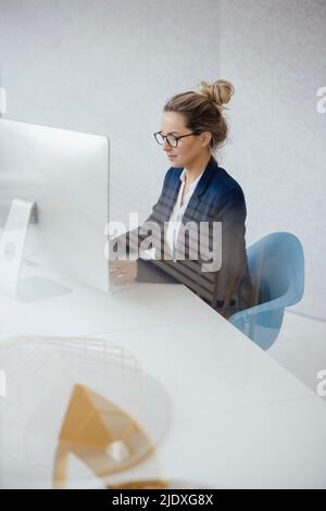 Donna d'affari che utilizza un PC desktop alla scrivania visto attraverso il vetro Foto Stock