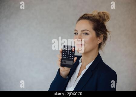 Donna d'affari sorridente che mostra il testo dell'asino sulla calcolatrice capovolta davanti al muro grigio Foto Stock