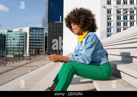 Giovane donna sorridente che indossa cuffie senza fili seduti con il telefono cellulare su gradini Foto Stock