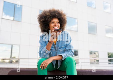 Donna sorridente che invia messaggi vocali tramite smartphone seduto sul banco Foto Stock