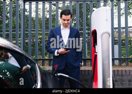 Uomo d'affari che utilizza uno smartphone in piedi presso la stazione di ricarica del veicolo elettrico Foto Stock