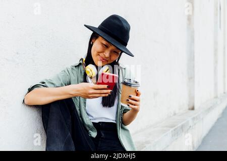 Donna sorridente con una tazza di caffè riutilizzabile rete da surf attraverso il telefono cellulare seduto a parete Foto Stock