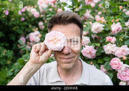 Uomo sorridente che tiene la rosa davanti all'occhio in giardino Foto Stock