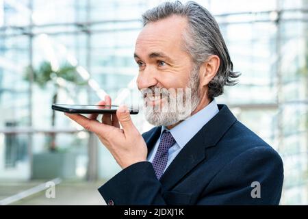 Uomo d'affari felice con capelli grigi che inviano messaggi vocali tramite smartphone Foto Stock
