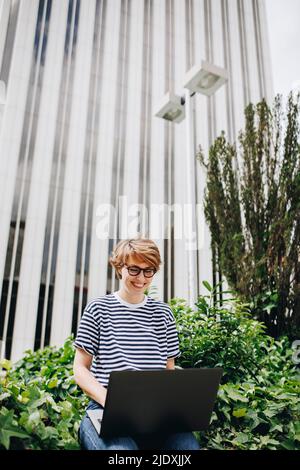 Donna che utilizza un computer portatile seduto di fronte all'edificio Foto Stock