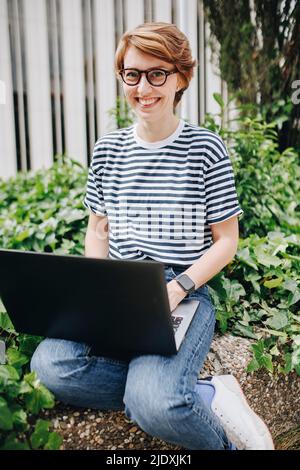 Donna felice con laptop seduto a parete da piante Foto Stock