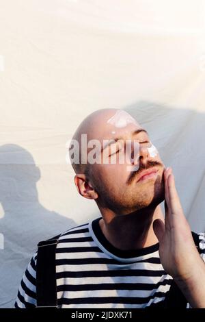 Calvo uomo con occhi chiusi applicando moisturizer di fronte a sfondo bianco Foto Stock