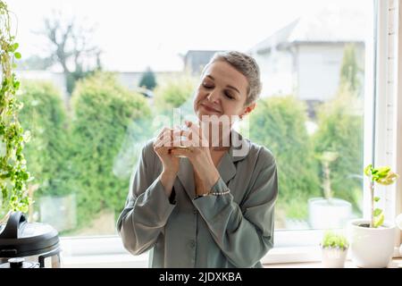 Donna matura che tiene il bicchiere di tè alla finestra a casa Foto Stock