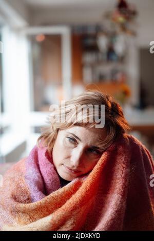 Donna matura pensierosa avvolta in coperta seduta a casa Foto Stock