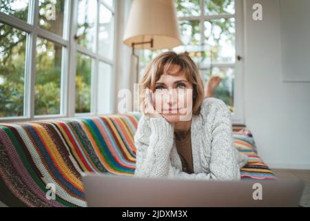 Donna matura e pensata con un computer portatile sul divano di casa Foto Stock