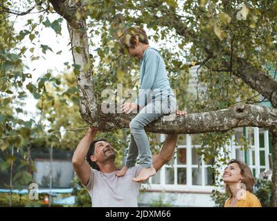 I genitori felici che guardano il figlio seduto sull'albero nel cortile posteriore Foto Stock