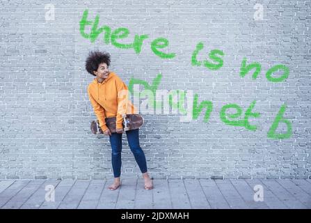 Donna che tiene lo skateboard di fronte al muro con testo non c'è Planet B. Foto Stock