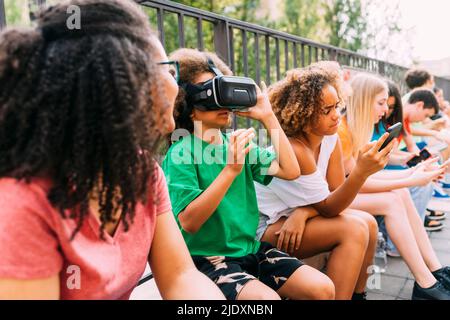 Ragazzo che indossa simulatore di realtà virtuale seduto con gli amici utilizzando lo smartphone Foto Stock