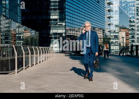 Uomo d'affari anziano che parla sullo smartphone che cammina all'esterno dell'edificio dell'ufficio Foto Stock