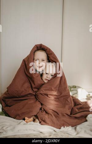 Fratelli sorridenti avvolti in coperta marrone a casa Foto Stock