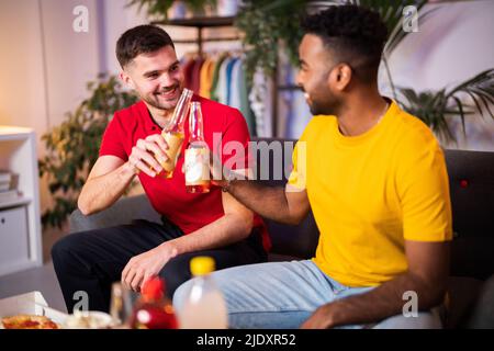 Giovani amici felici che tostano bottiglie di birra seduti a casa Foto Stock