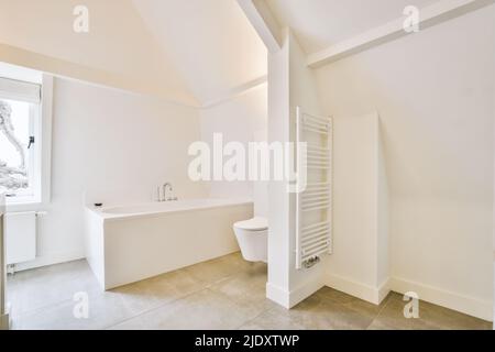 Vasca bianca con rubinetto in metallo accanto alla finestra e wc sospeso in un moderno bagno in stile minimalista nell'appartamento Foto Stock