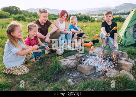 Sei bambini gruppo ragazzi e ragazze allegramente ridero e arrostite salsicce su bastoni sopra una fiamma fuoco vicino alla tenda verde Foto Stock