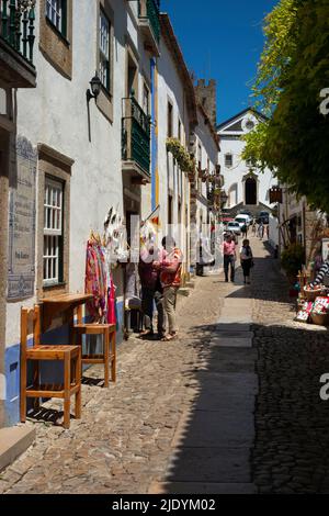 I turisti curiosano tra i souvenir di Rua Direita a Óbidos, nella regione del Centro in Portogallo, sotto l'ex Igreja de Santiago o São Tiago, la fine del 18th secolo ex Chiesa di San Giacomo, che ora ospita la libreria Livraria de Santiago. Foto Stock