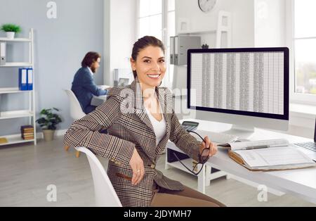 Ritratto di felice sorridente contabile finanziaria seduta alla scrivania d'ufficio con il computer Foto Stock