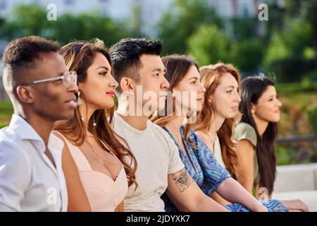 Gruppo di giovani multirazziali che guardano via mentre si siedono in fila insieme all'aperto. Concetto di squadra e unità. Foto Stock