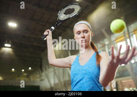 Giocatore di tennis femminile di grande fiducia con treccia in piedi sul campo interno e lanciare la palla mentre si pratica il colpo Foto Stock