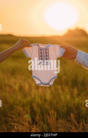 Le mani di uomo e donna tengono il vestito neonato con ricamo nazionale ucraino sullo sfondo del tramonto sul prato. Concetto di attesa del bambino. Foto Stock