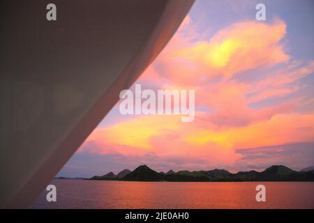 Il cielo colorato, le nuvole sopra l'acqua di mare e il paesaggio collinare intorno al tramonto sono visti da una barca all'interno dell'area del Parco Nazionale di Komodo a Komodo, Manggarai Occidentale, Nusa Tenggara Orientale, Indonesia. Foto Stock