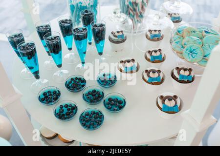Serve una fila di bicchieri di rinfrescanti bevande colorate con mirtillo, torte e caramelle, sfondo bianco. Buffet in stile blu, celebrazione dell'evento Foto Stock