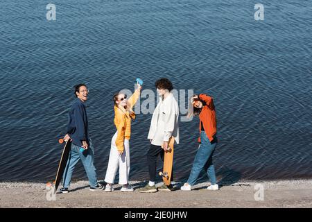 intera lunghezza di donna felice con lattina di soda in mano rialzata vicino amici multietnici sulla riva del fiume Foto Stock