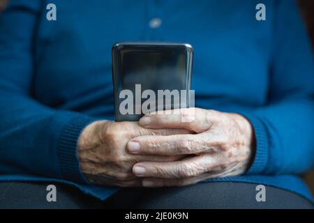 Le mani della donna anziana tengono un telefono cellulare e guardano lo schermo. Foto Stock