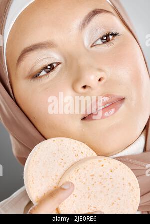 Bella donna musulmana che tiene e usando le spugne del trucco sul suo viso contro uno sfondo grigio studio. Giovane donna che indossa un hijab e la fa Foto Stock