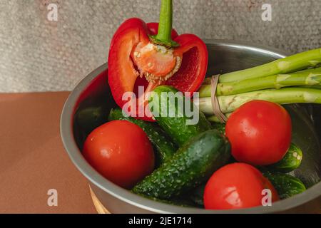 Le verdure si trovano in una ciotola di metallo: Pomodori, asparagi, cetrioli, peperoni rossi . su un asse di legno e sfondo marrone. Sfondo grigio posteriore. Luogo Foto Stock