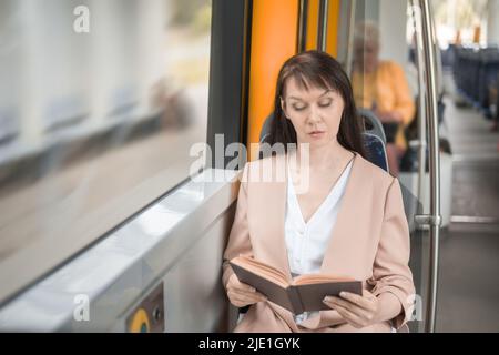 una donna adulta si siede in un tram e legge un interessante libro durante il viaggio. Foto Stock