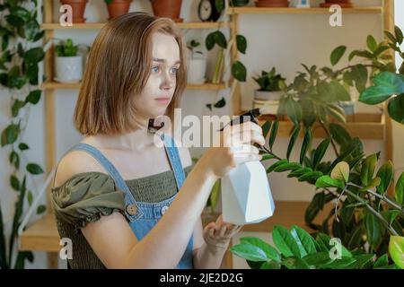 Una giovane donna si prende cura delle sue piante di casa teneramente spruzzandole. Sta osservando la malattia, preoccupandosi delle sue piante. Giardinaggio domestico, concetto di hobby. Foto Stock