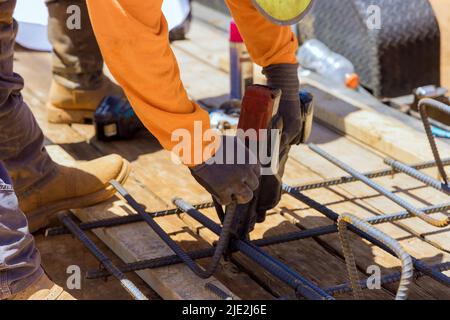 Le mani dei lavoratori edili fissano le barre in acciaio con le aste metalliche per rinforzare il cemento utilizzando l'attrezzo di legatura delle barre di rinforzo Foto Stock